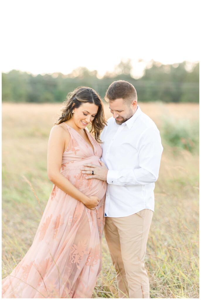 A pregnant mother and her husband holding their pregnant belly outside in a field looking down at the belly while taking their maternity photos in DC